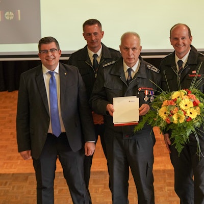 Gruppenbild mehrere Männer. Ein Feuerwehrmann hält eine Urkunde in der Hand. Ein weiterer Feuerwehrmann neben ihm einen Blumenstrauß.