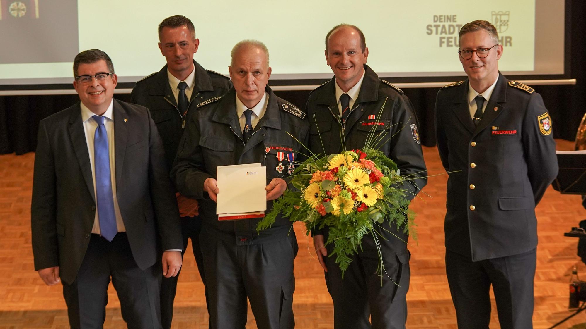 Gruppenbild mehrere Männer. Ein Feuerwehrmann hält eine Urkunde in der Hand. Ein weiterer Feuerwehrmann neben ihm einen Blumenstrauß.