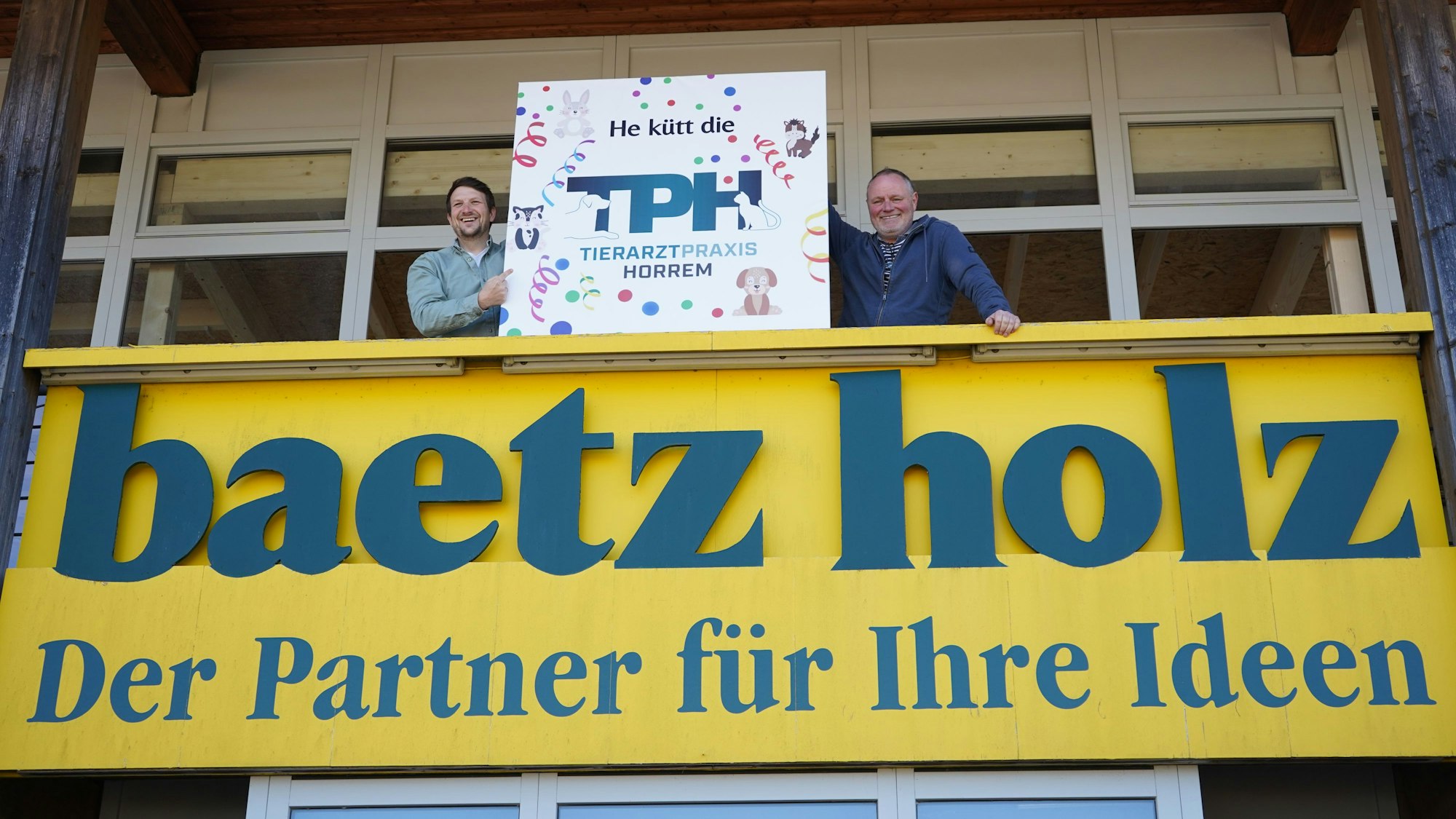 Das Foto zeigt Ludwig Münch und Boris Burghardt auf einem Balkon, auf dem der Schriftzug „baetz holz Der Partner für Ihre Ideen“ steht. Sie halten ein Schild hoch, auf dem „Tierarztpraxis Horrem“ steht.