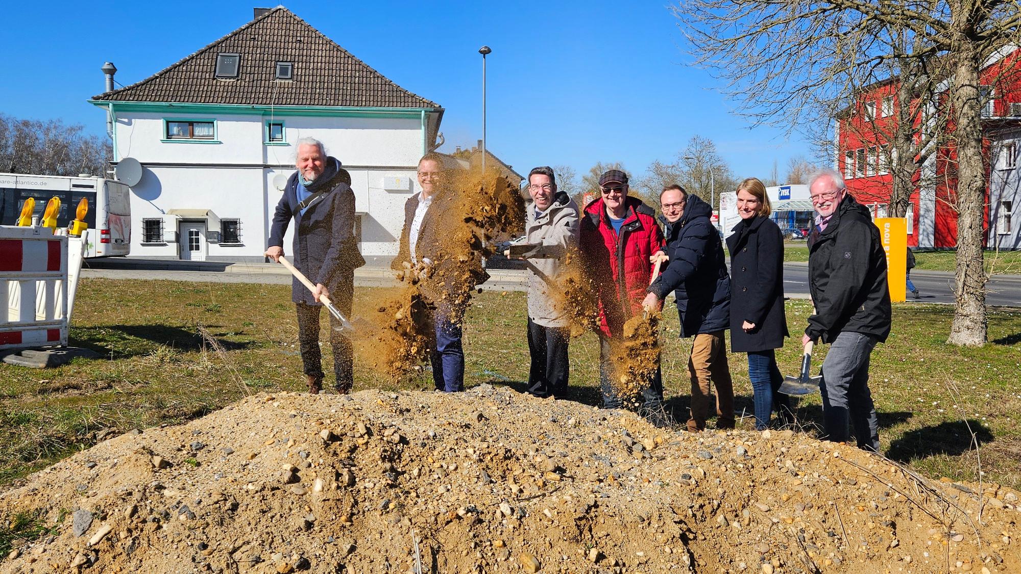 Zu sehen sind mehrere Männer und eine Frau, die mit Spaten an einer Baustelle Erde werfen.
