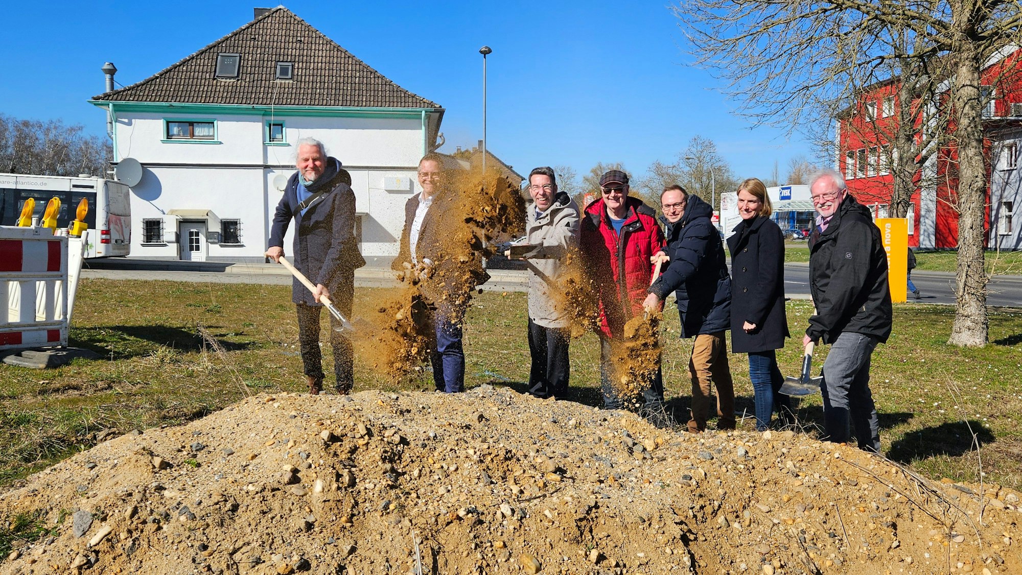 Zu sehen sind mehrere Männer und eine Frau, die mit Spaten an einer Baustelle Erde werfen.