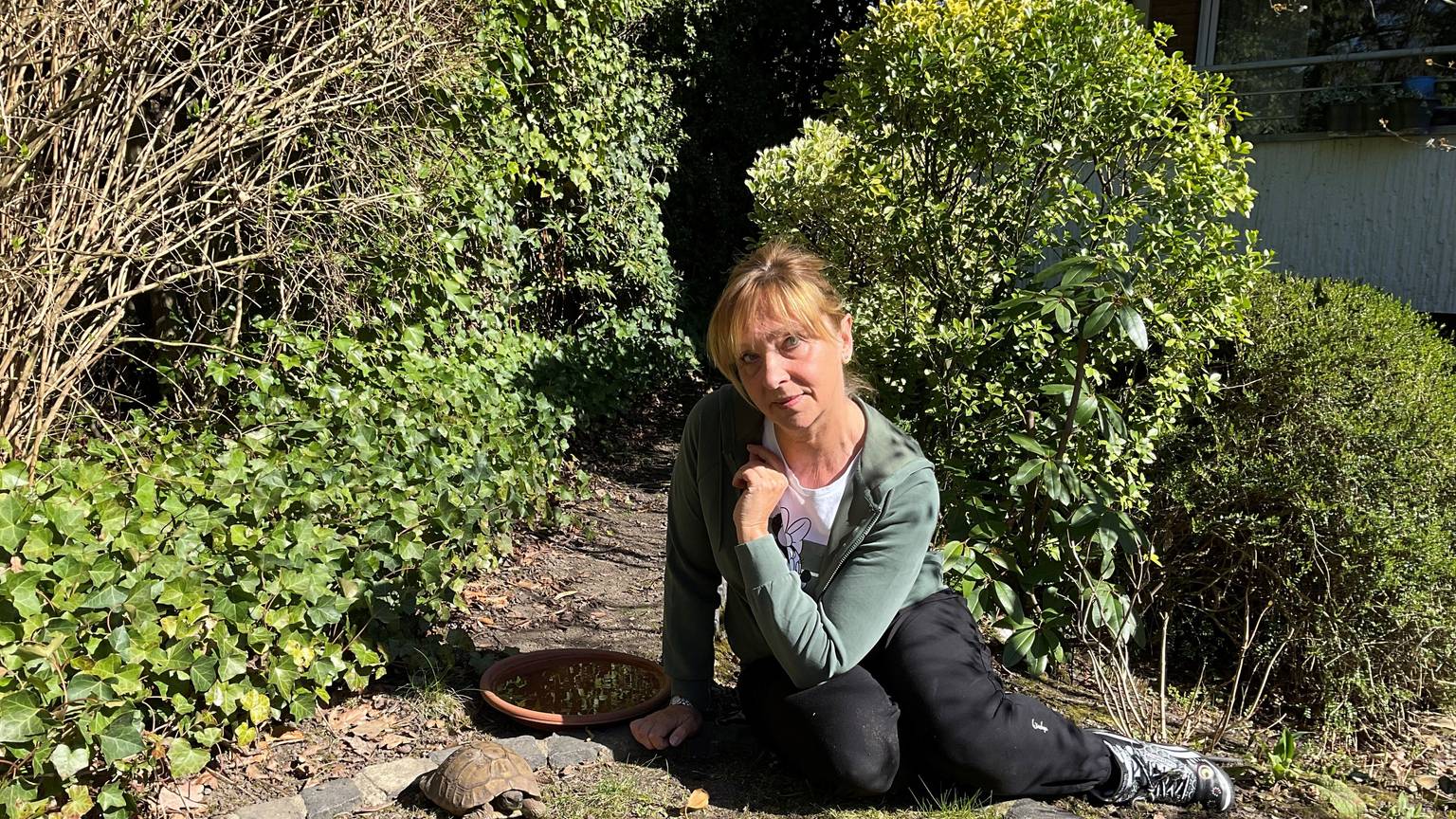 Eine Frau sitzt neben einer Landschildkröte in einem Garten.