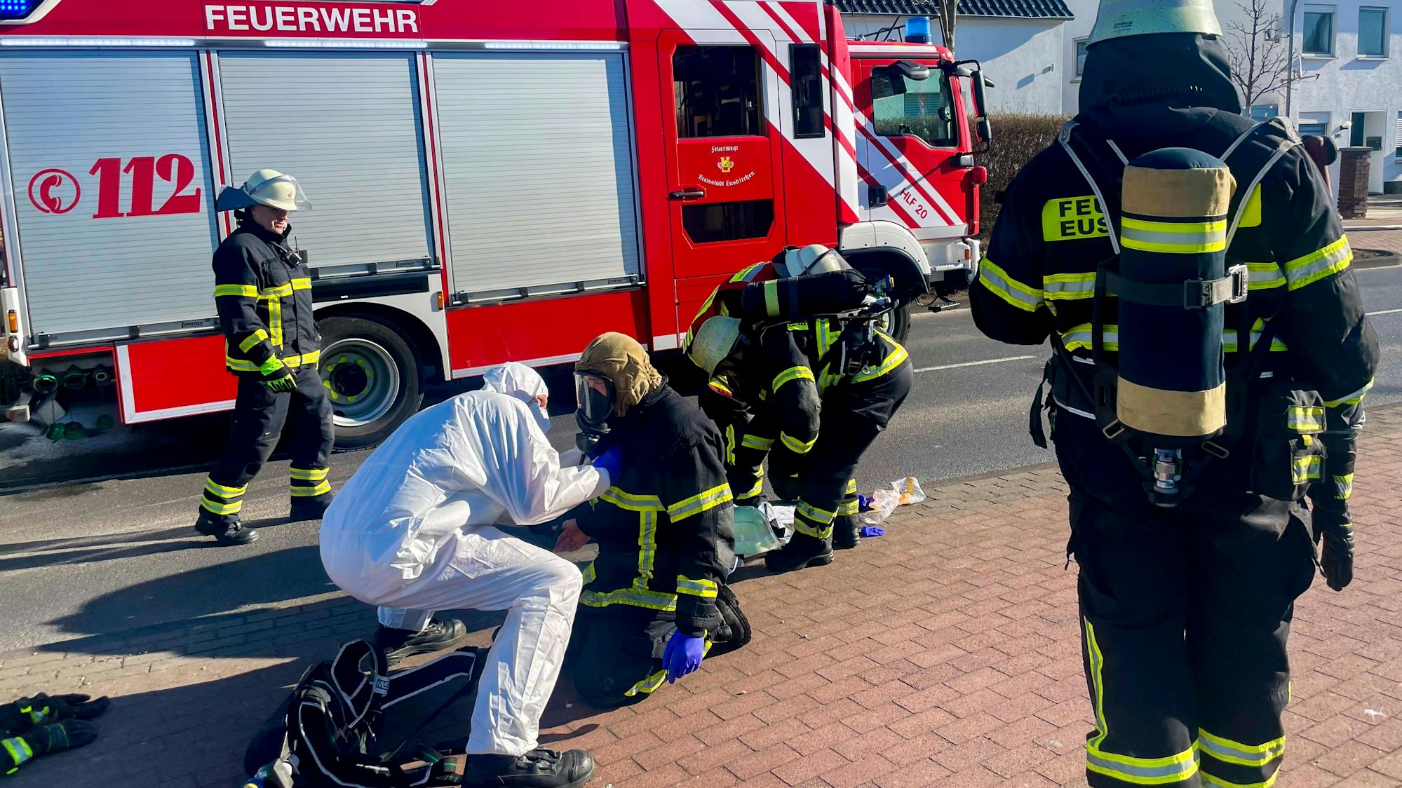 Das Bild zeigt einen Feuerwehrmann, der auf dem Bürgersteig kniet. Ein Mann in einem weißen Schutzanzug öffnet ihm die Jacke.