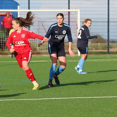 Saskia Reder führt den Ball, die Zunge hat sie in die Wange gepresst, eine Gegenspielerin mit blutendem Knie schaut zu.