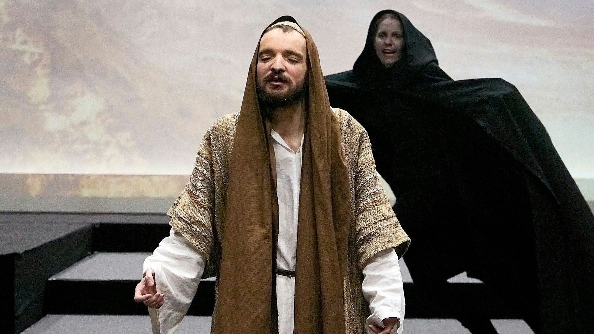 Der Jesus-Darsteller in einem hellen Gewand mit braunem Schal hat die Augen geschlossen. Hinter ihm breitet eine schwarz gekleidete Frau in der Rolle des Satans ihre Arme aus, dahinter eine projizierte Wüstenkulisse.