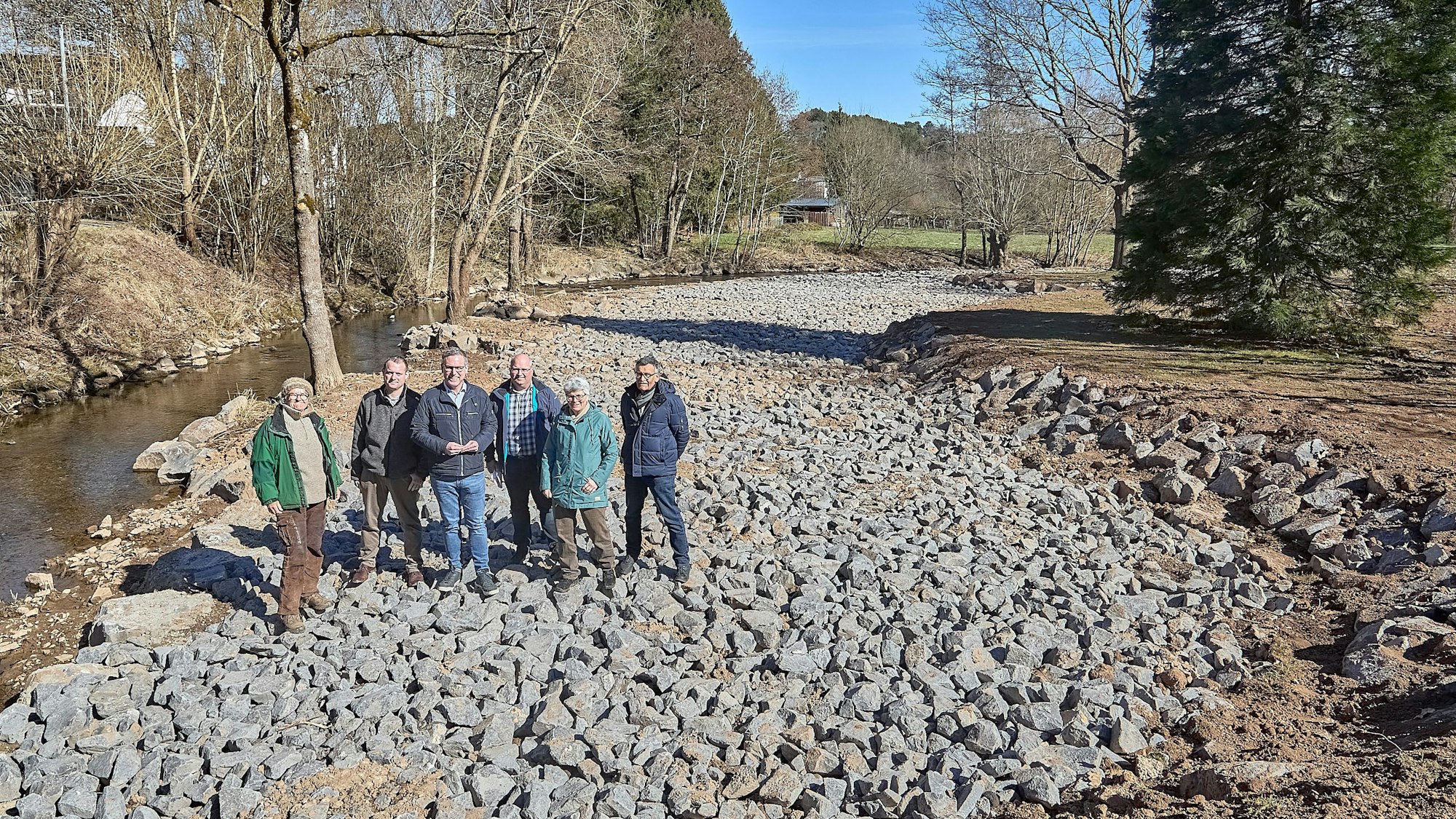 Sechs Personen stehen an einem Fluss auf einer Schotterfläche.