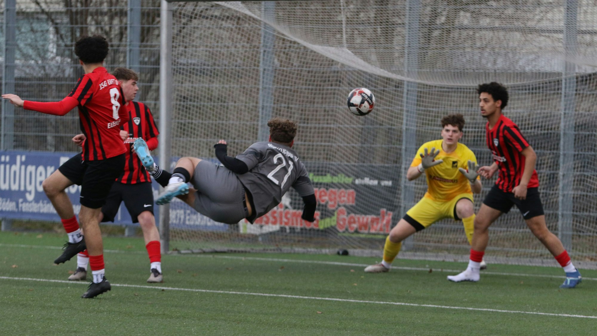 Ein Fußballspieler versucht per Fallrückzieher ein Tor zu erzielen. Den Ball wehrt ein Verteidiger noch vor dem Torwart ab.