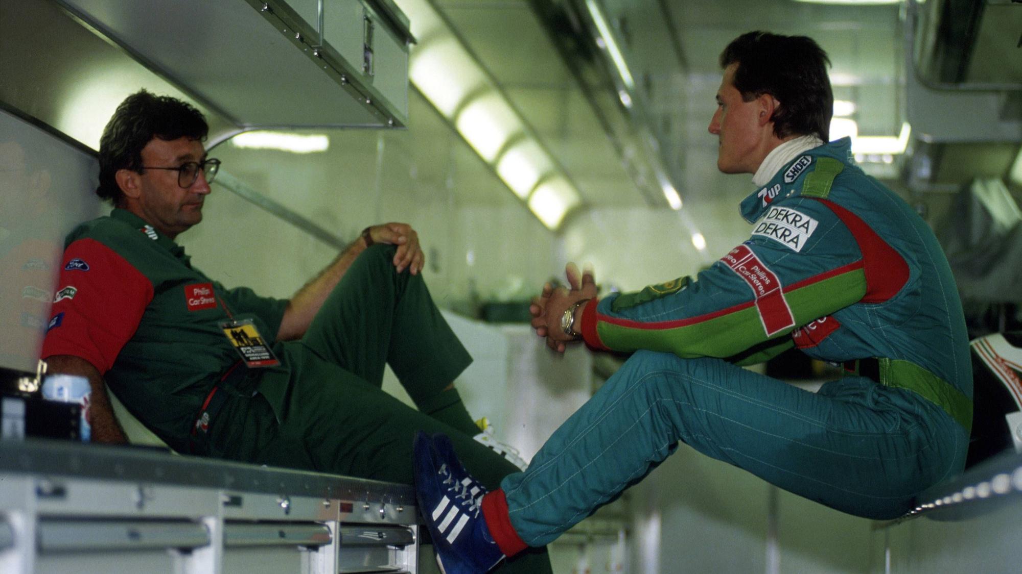 Michael Schumacher im Gespräch mit Teamchef Eddie Jordan. Die beiden sitzen sich gegenüber. Schumacher trägt einen blauen Rennanzug.