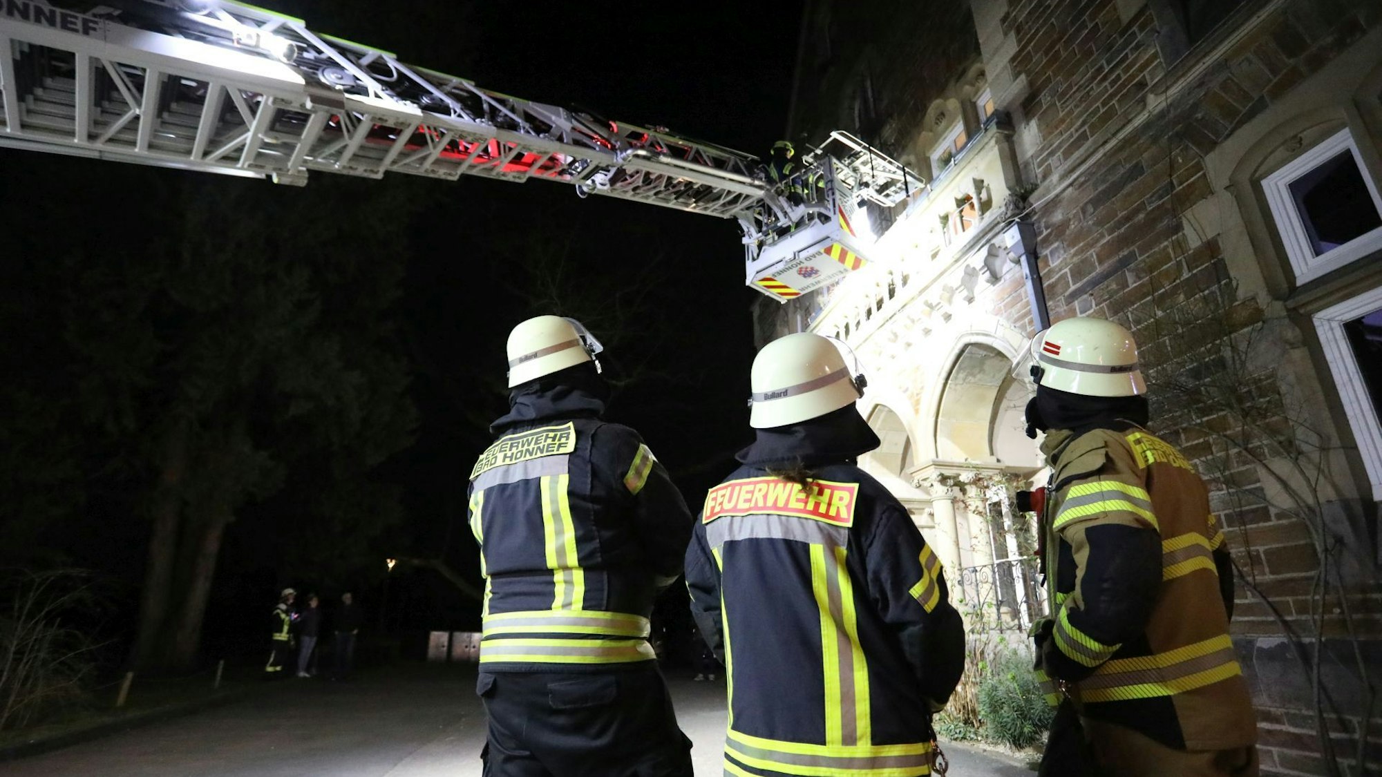 Drei Feuerwehrleute beobachten den Einsatz einer Drehleiter am Balkon eines historischen Gebäudes.
