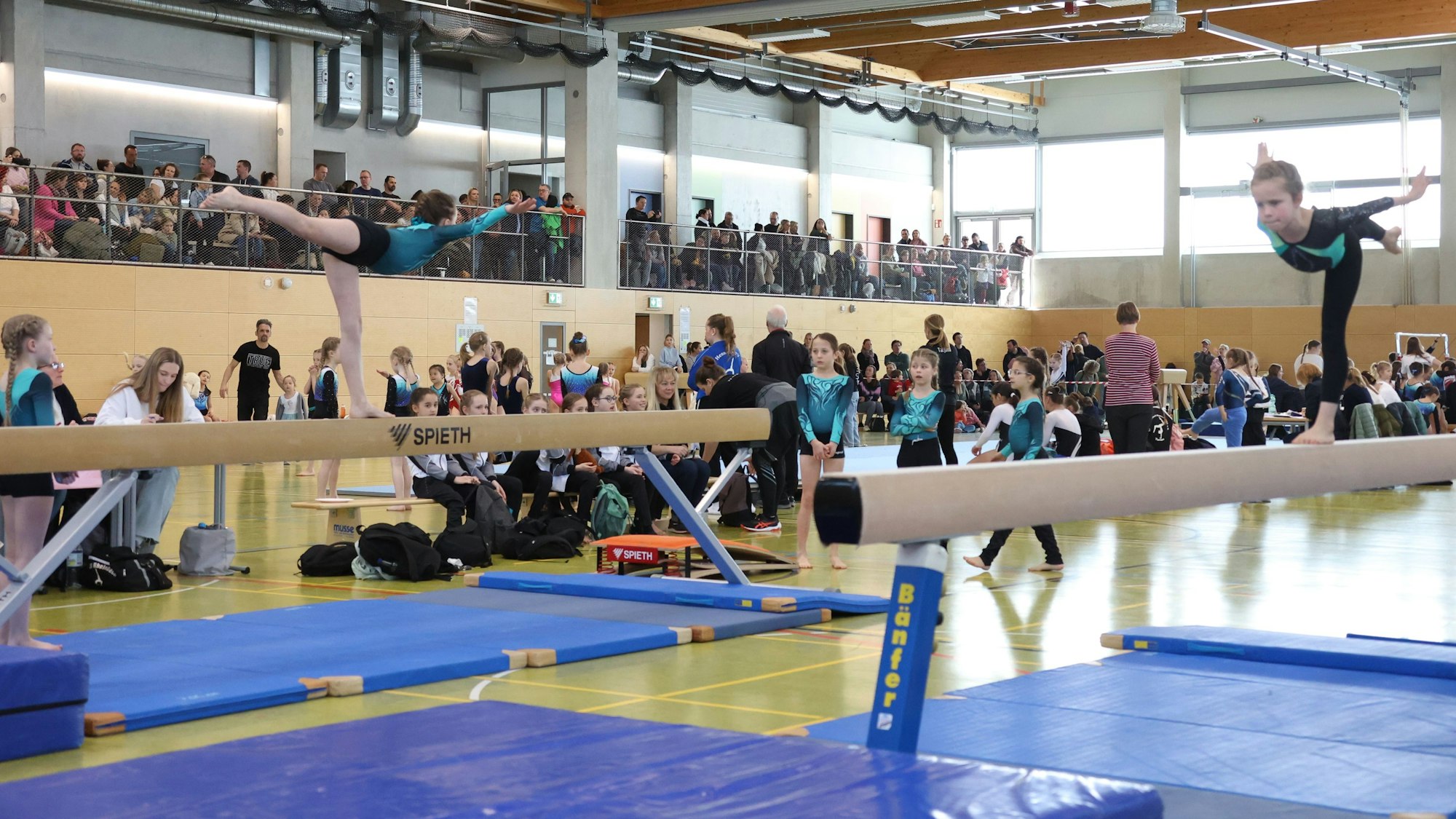 Auf zwei Schwebebalken turnen junge Mädchen. Im Hintergrund sind zahlreiche Zuschauer zu erkennen.