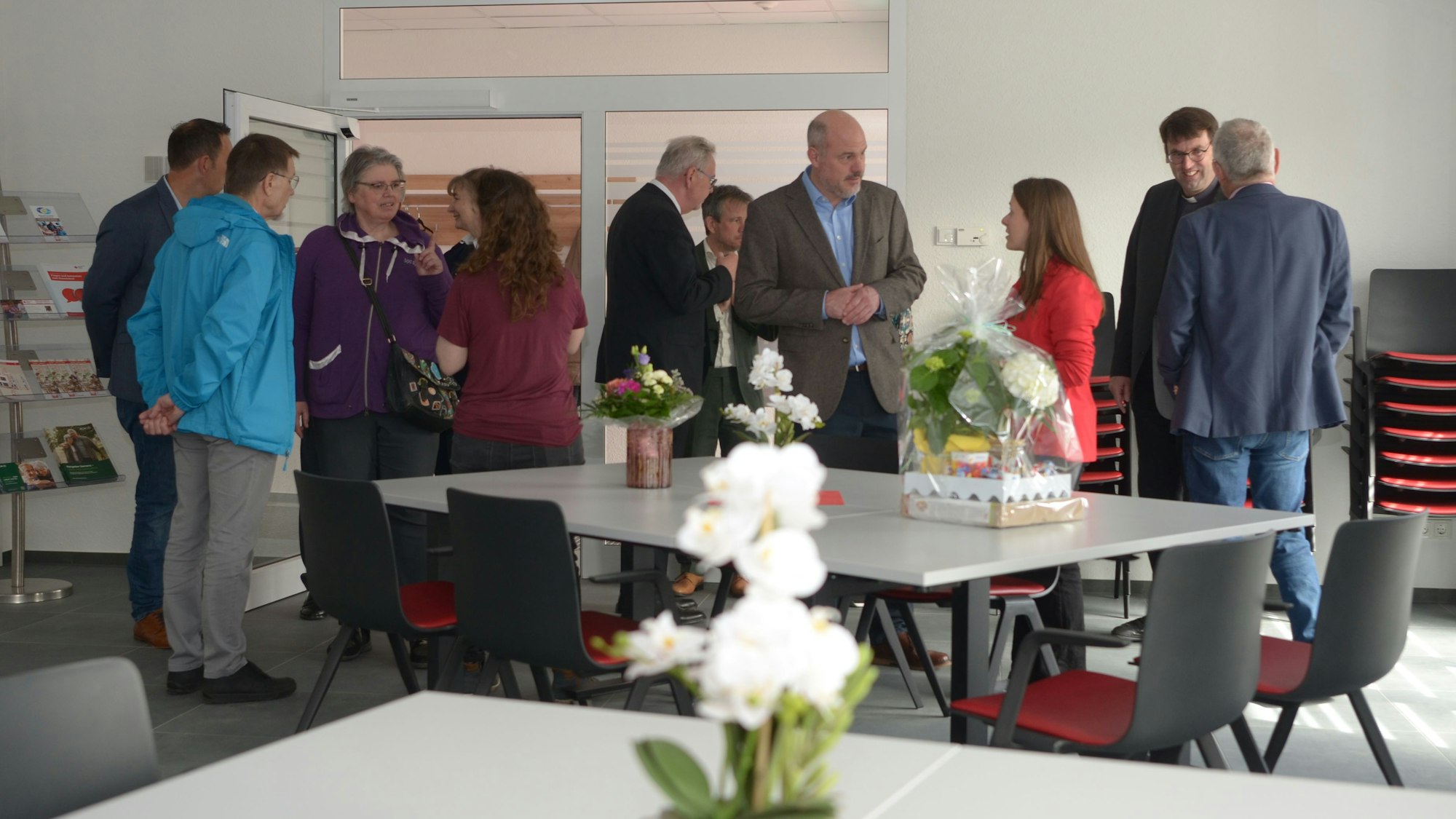 Das Foto zeigt den Raum mit Tischen, Stühlen und Blumen. Im Hintergrund unterhalten sich Besucherinnen und Besucher.