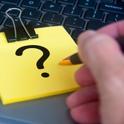 Ein gelber Notizblock, auf dessen oberstem Blatt ein schwarzes Fragezeichen zu sehen ist, liegt auf einem aufgeklappten Laptop.