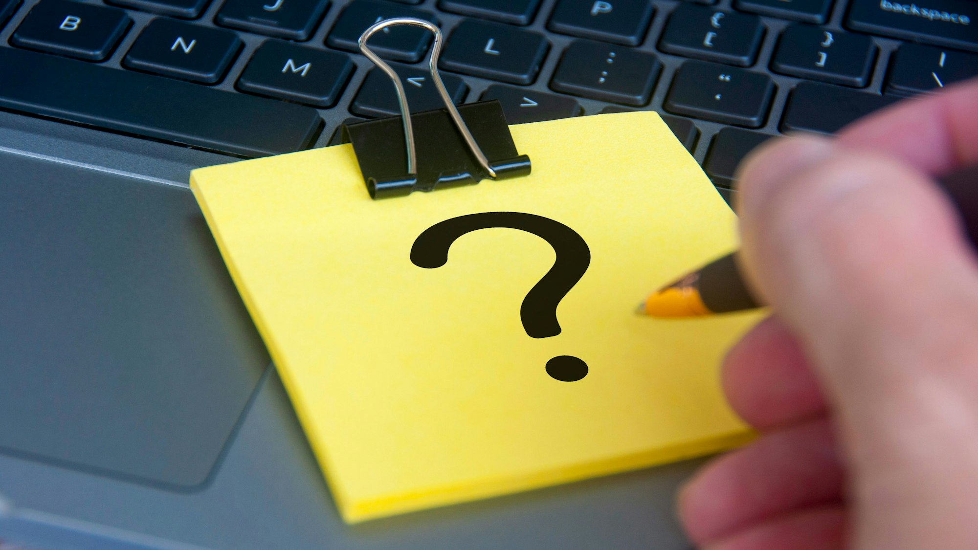 Ein gelber Notizblock, auf dessen oberstem Blatt ein schwarzes Fragezeichen zu sehen ist, liegt auf einem aufgeklappten Laptop.