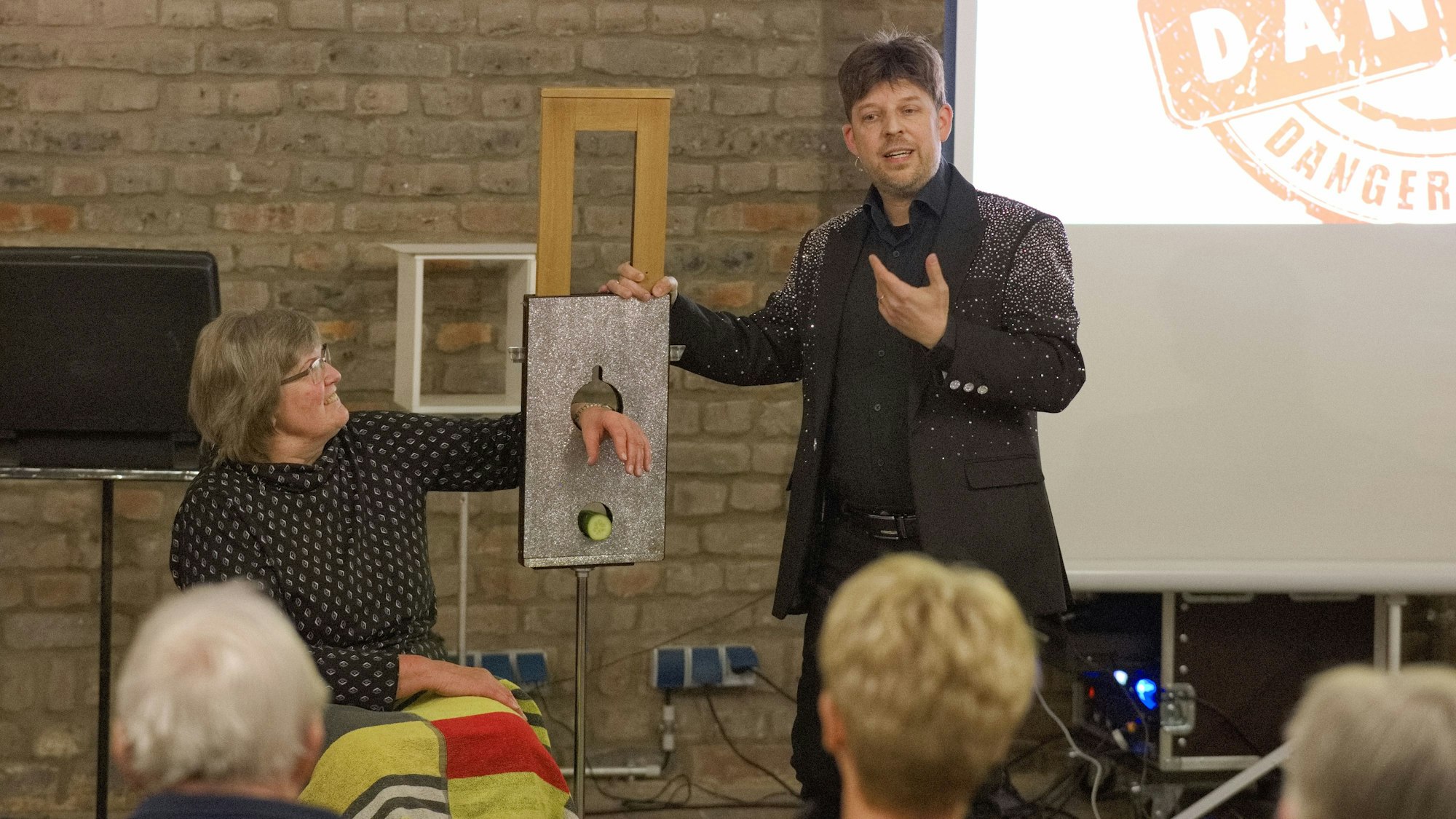 Der Magier Carsten Fenner erklärt dem Publikum im Kulturhof Velbrück, warum der Trick mit der Guillotine meistens gut ausgeht. Eine Zuschauerin, die ihre Hand in das Loch unter dem Fallmesser gelegt hat, schaut skeptisch.