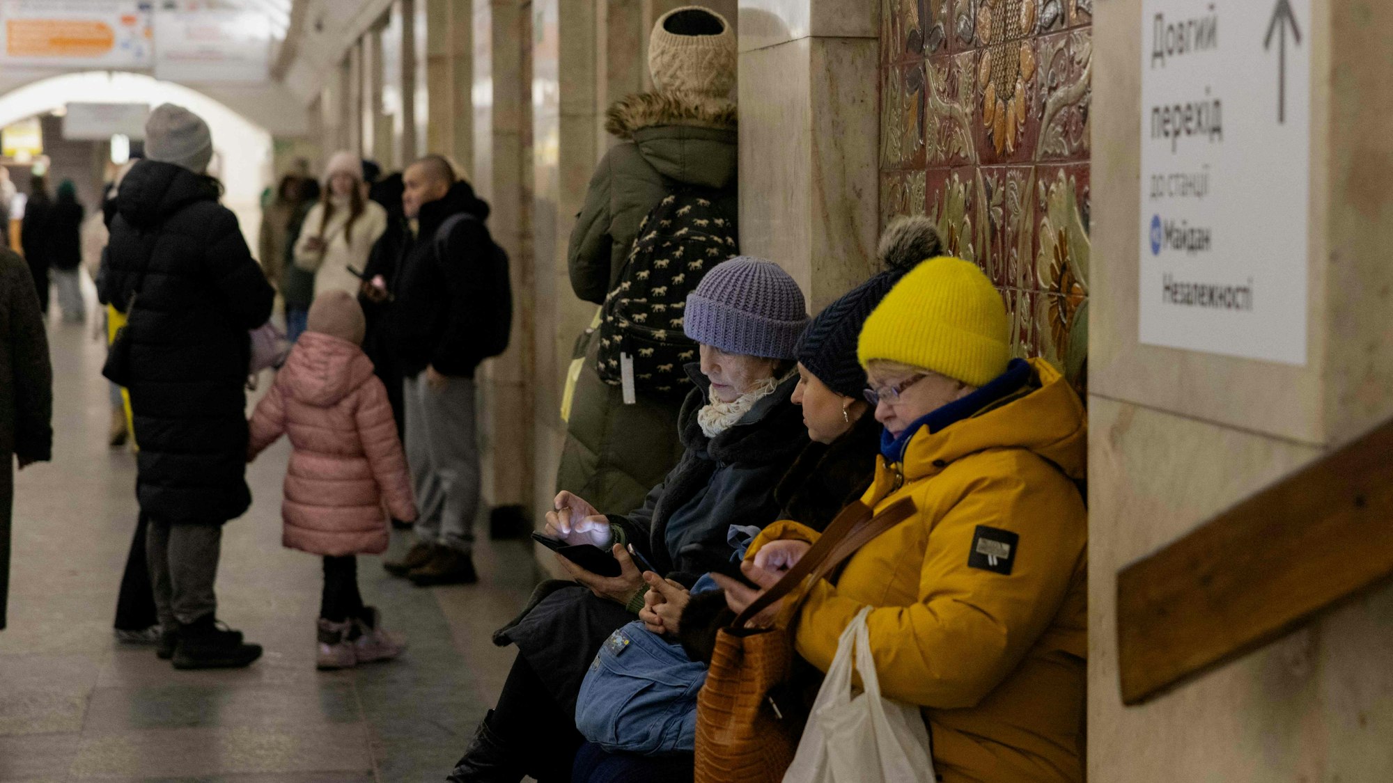 Kiewerinnen sitzen in Winterjacken auf einer Bank in einer U-Bahn-Station.