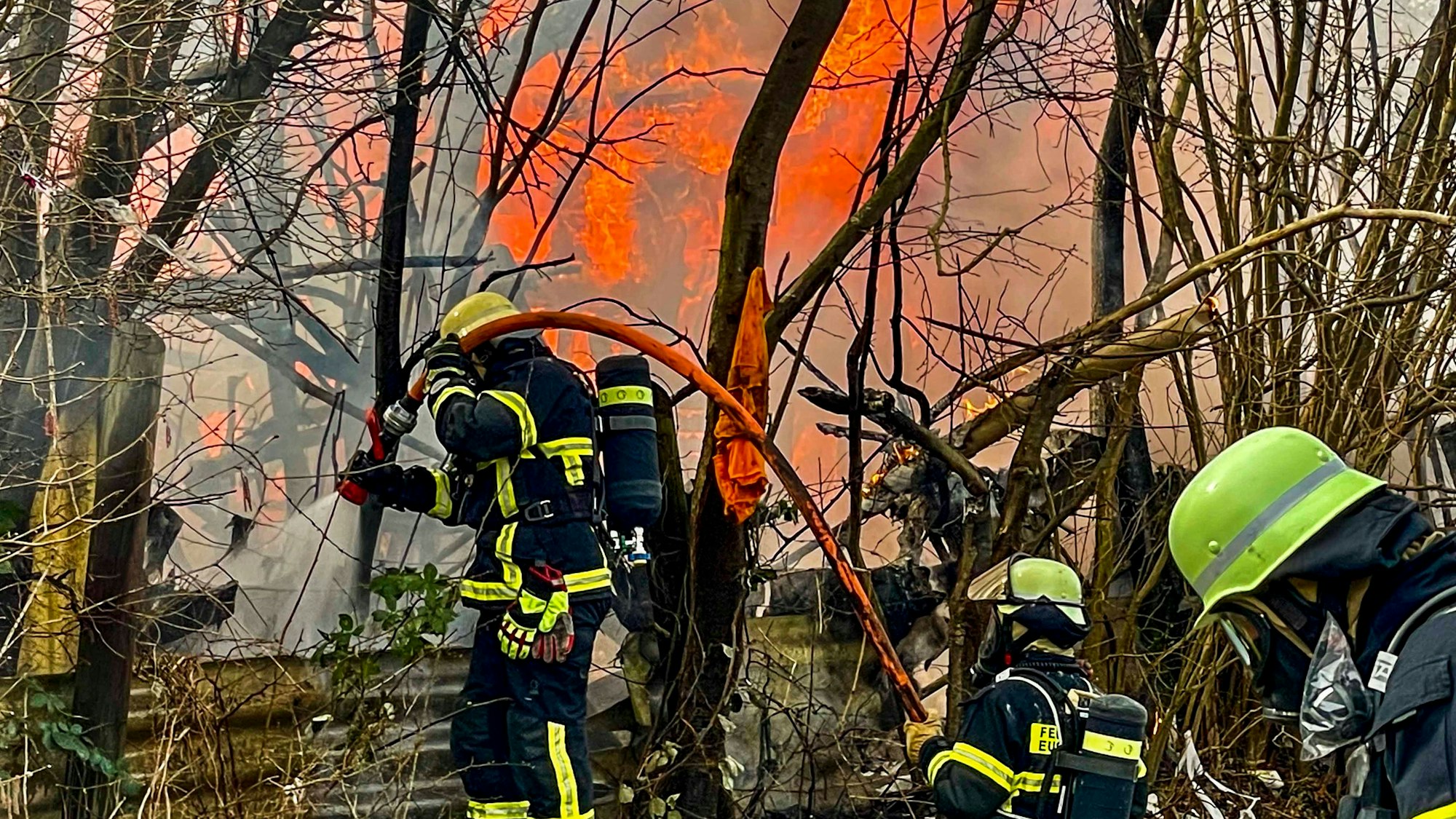 Das Bild zeigt drei Feuerwehrleute, die den Brand bekämpfen. Im Hintergrund sind Flammen zu sehen.