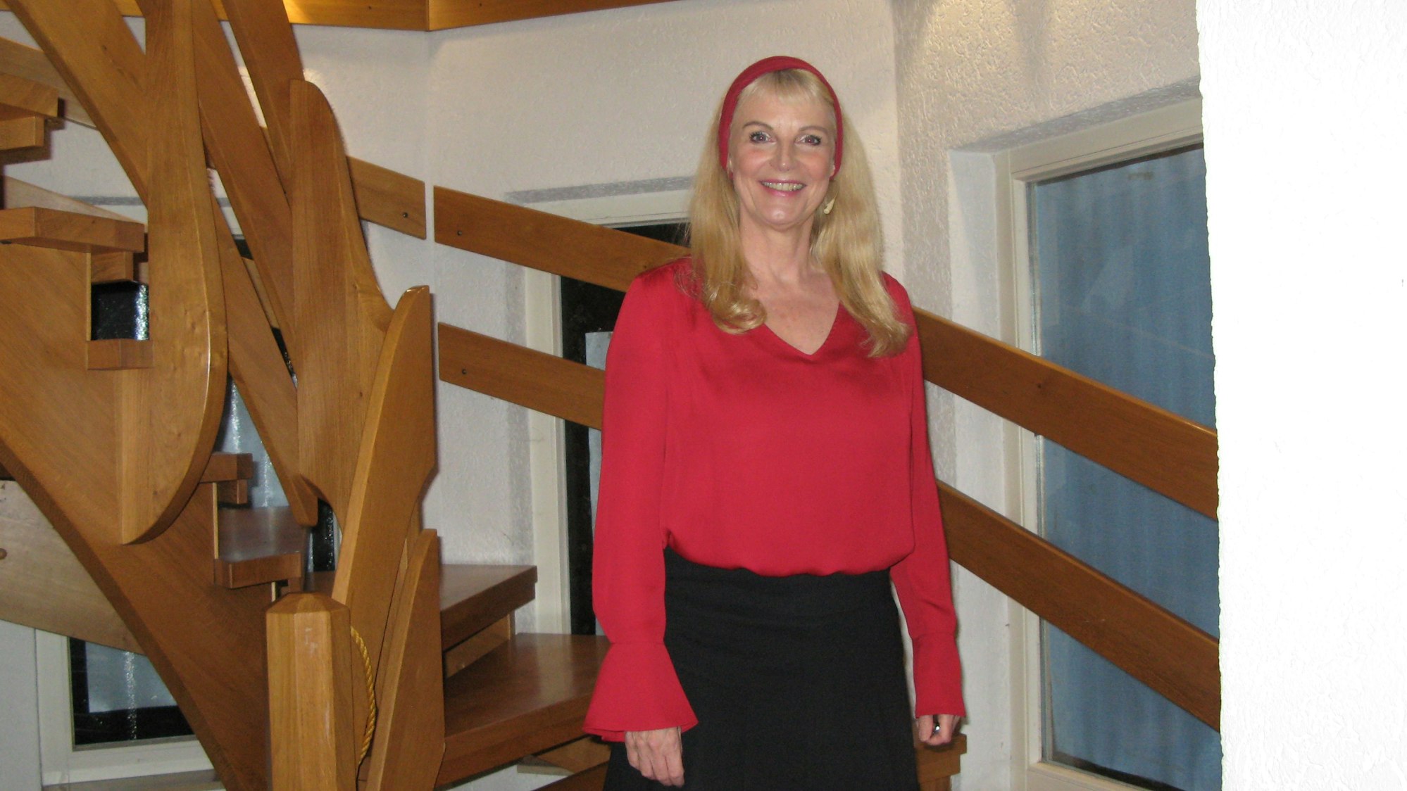 Eine Frau mit langen blonden Haaren, roter Bluse und schwarzem Rock steht auf einer Treppe und lächelt.