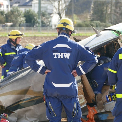 Männer in Overalls des THW stehen vor dem Wrack des abgestürzten Flugzeuges am Flugplatz in Sankt Augustin.