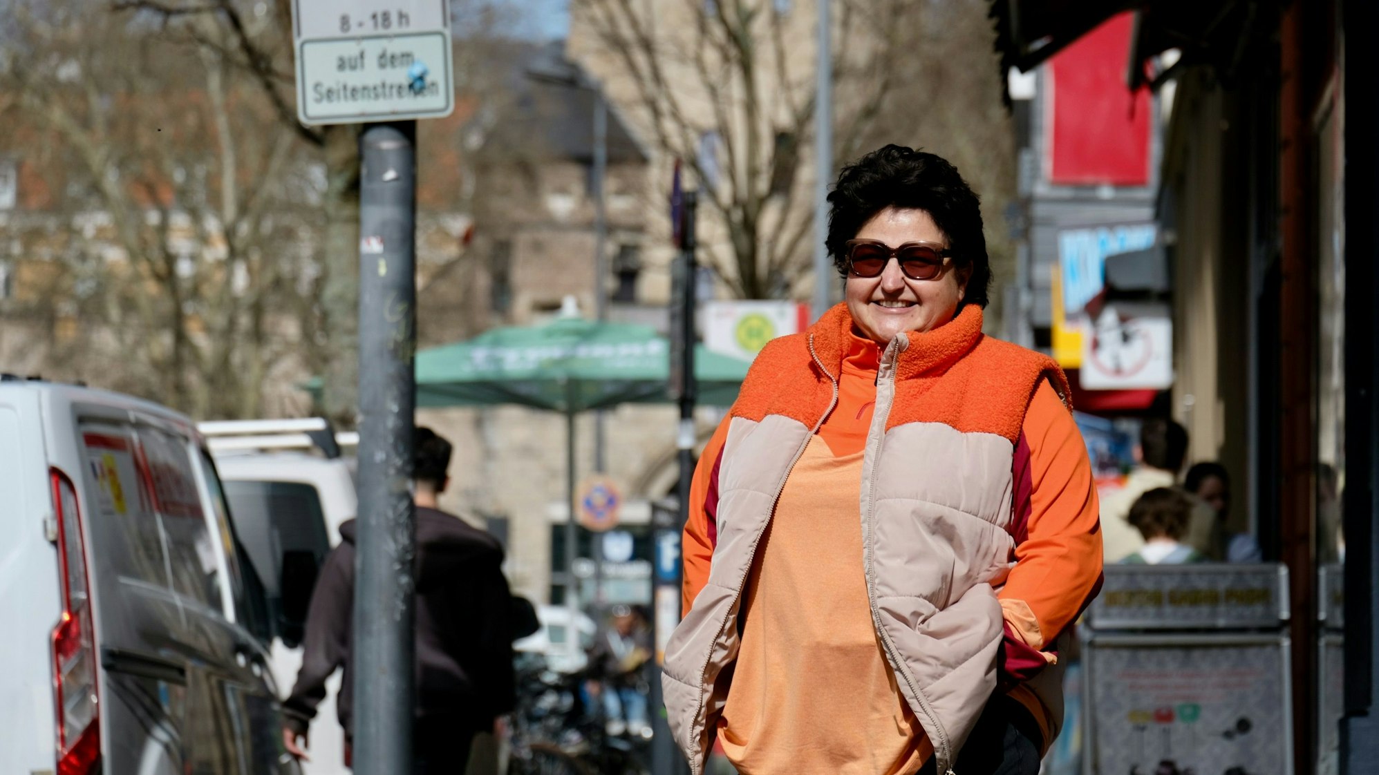Eine Frau steht auf einer belebten Straße im Sonnenschein, sie trägt eine Sonnenbrille.