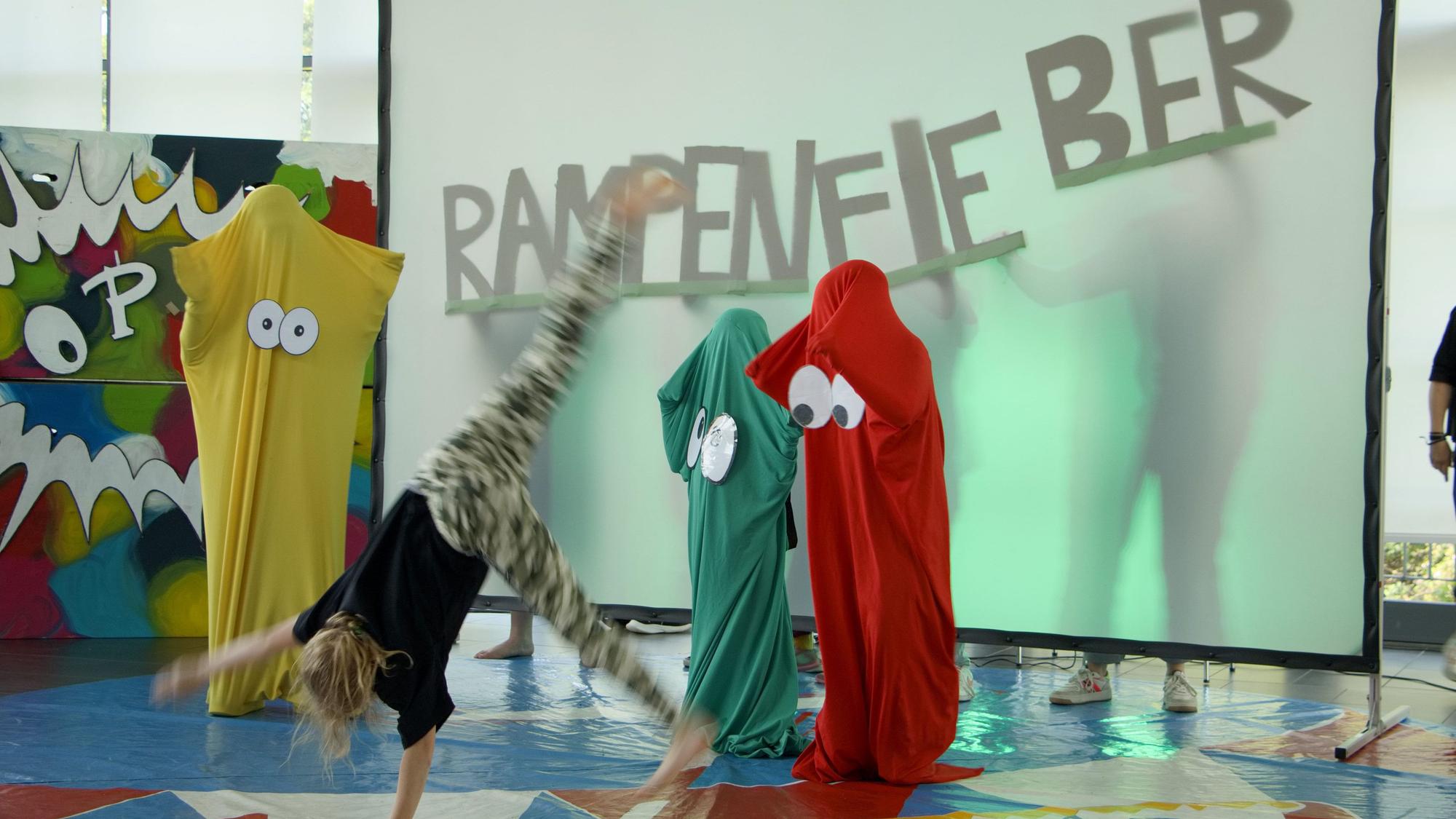 Ein Kind schlägt auf einer Bühne ein Rad. Im Hintergrund steht auf einer Leinwand das Wort „Rampenfieber“.