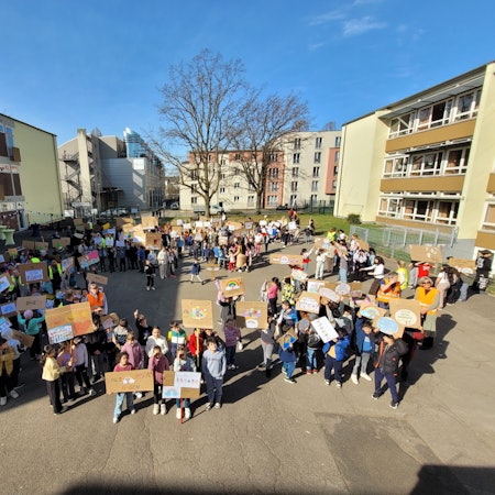 Zahlreiche Schulkinder stehen auf einem Schulhof und halten selbsterstellte Plakate in die Höhe.
