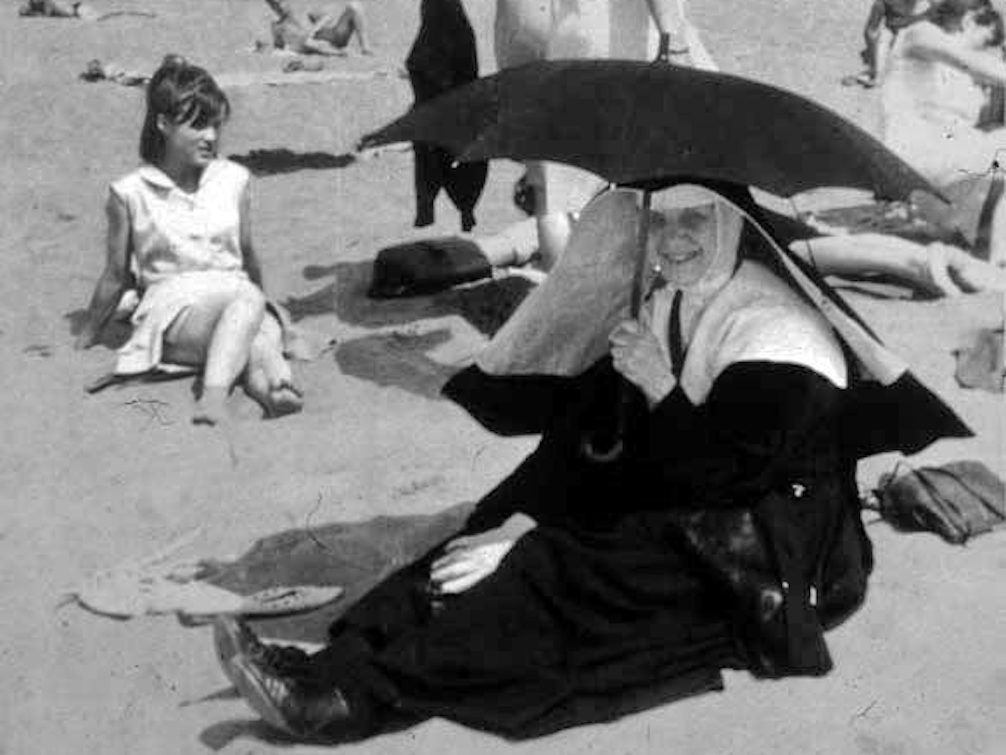 Eine Nonne sitzt im Sand an einem Strand, sie hält einen Regenschirm, um sich vor der Sonne zu schützen.