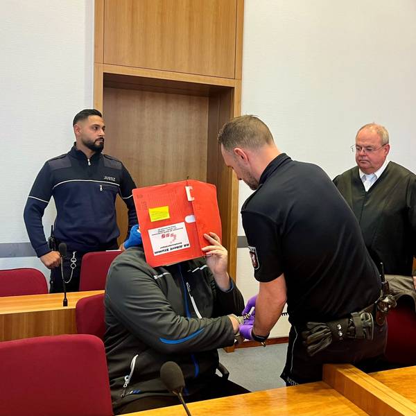 Das Foto zeigt den Angeklagten, der sein Gesicht mit einer Kladde verdeckt. Ein Justizwachtmeister löst ihm die Handfesseln, der Verteidiger beobachtet die Szene.