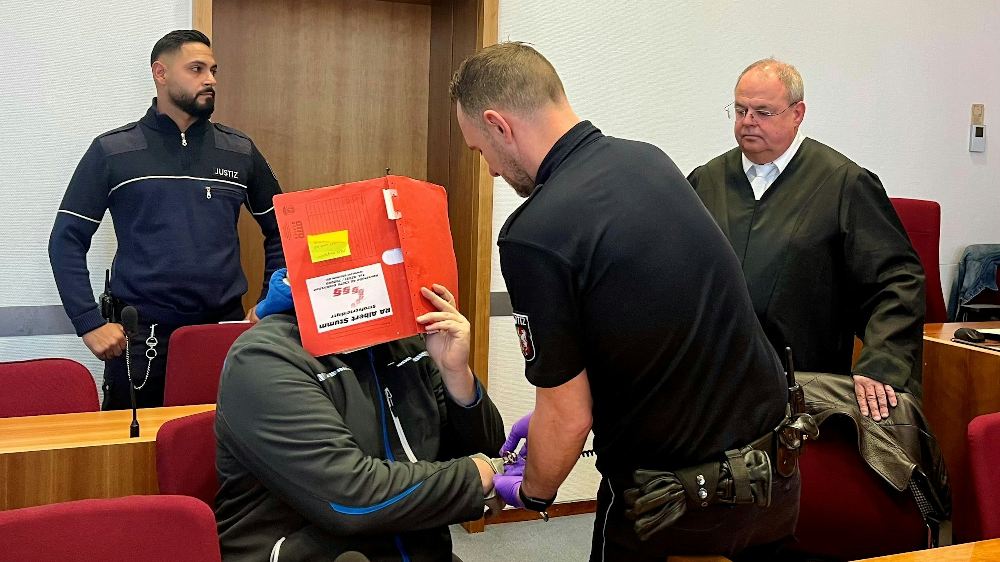 Das Foto zeigt den Angeklagten, der sein Gesicht mit einer Kladde verdeckt. Ein Justizwachtmeister löst ihm die Handfesseln, der Verteidiger beobachtet die Szene.