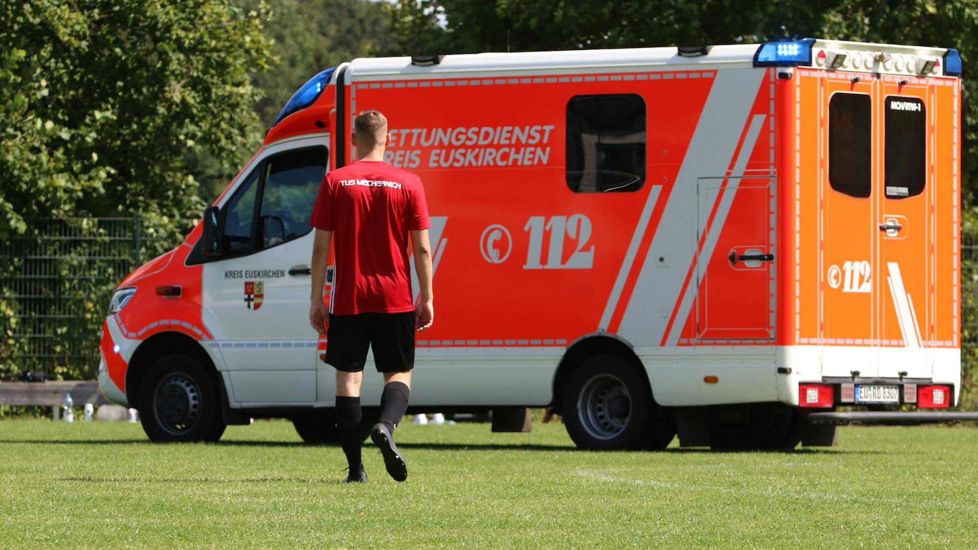 Ein Fußballer trottet in Richtung eines Rettungswagens, der auf dem Rasen eines Sportplatzes steht.
