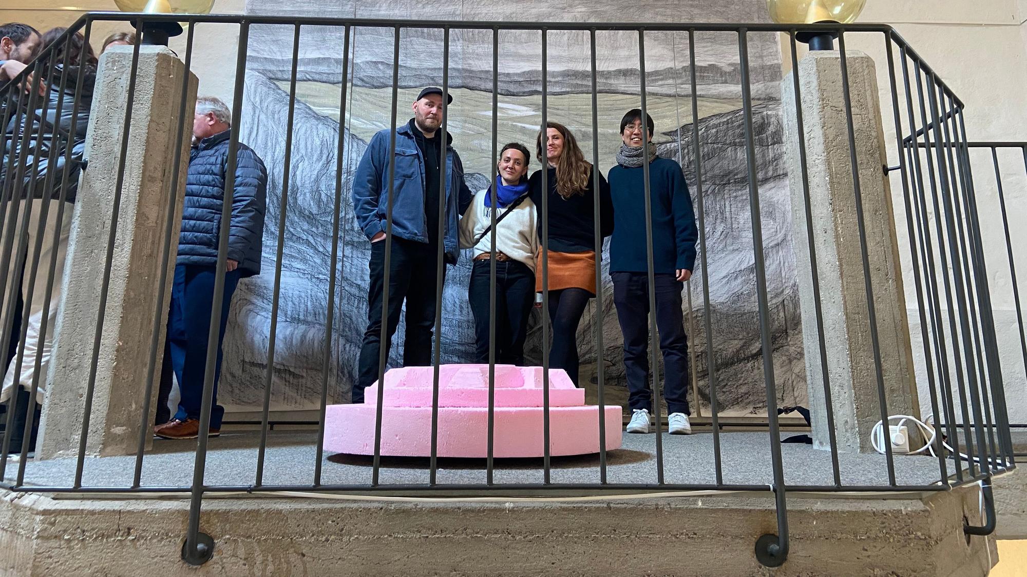 Vier Menschen stehen vor einem großen Gemälde, sie wurden durch ein Treppengeländer hindurch fotografiert.