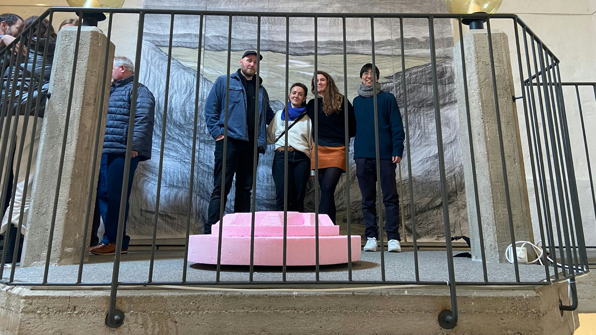 Vier Menschen stehen vor einem großen Gemälde, sie wurden durch ein Treppengeländer hindurch fotografiert.