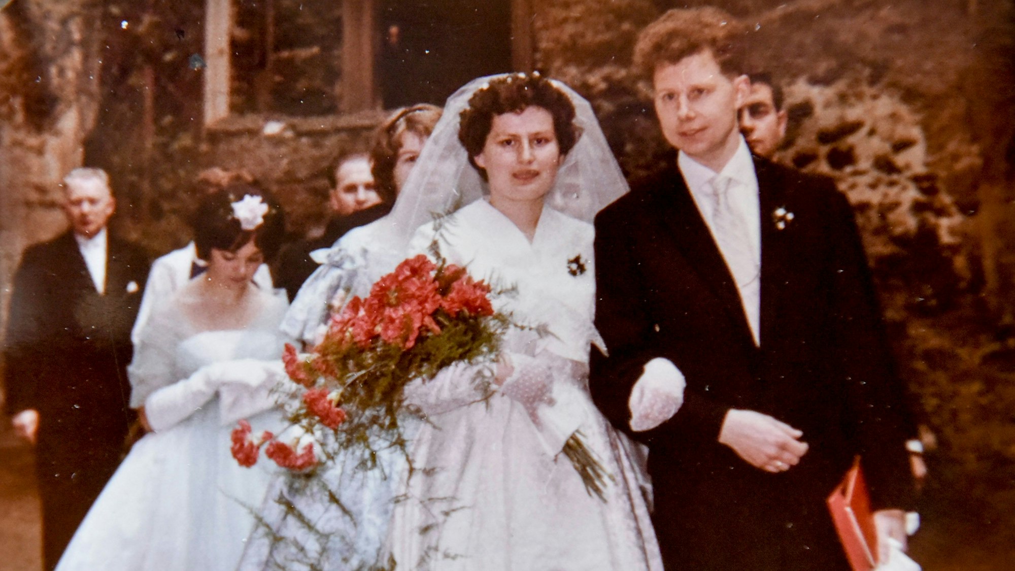 Ein Bräutigam führt seine Braut mit rotem Blumenstrauß und weißem Kleid zum Altar.