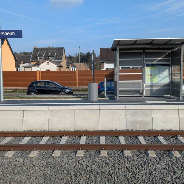 Das Bild zeigt einen Bahnsteig, auf dem sich ein Wartehäuschen, eine Bank und ein Mülleimer befinden.