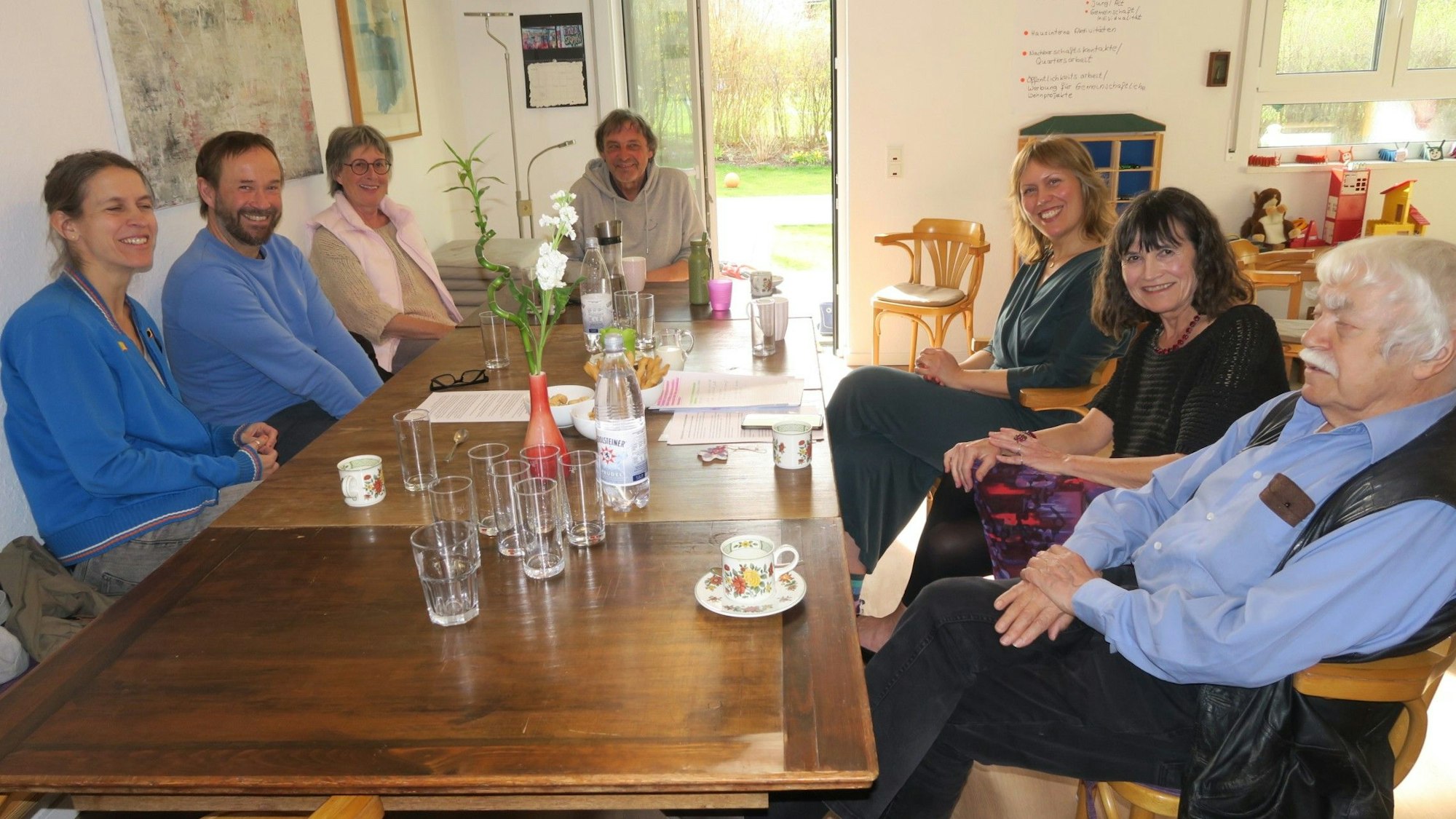 Sieben Personen, von Jung bis Alt, sitzen an einem Küchentisch.