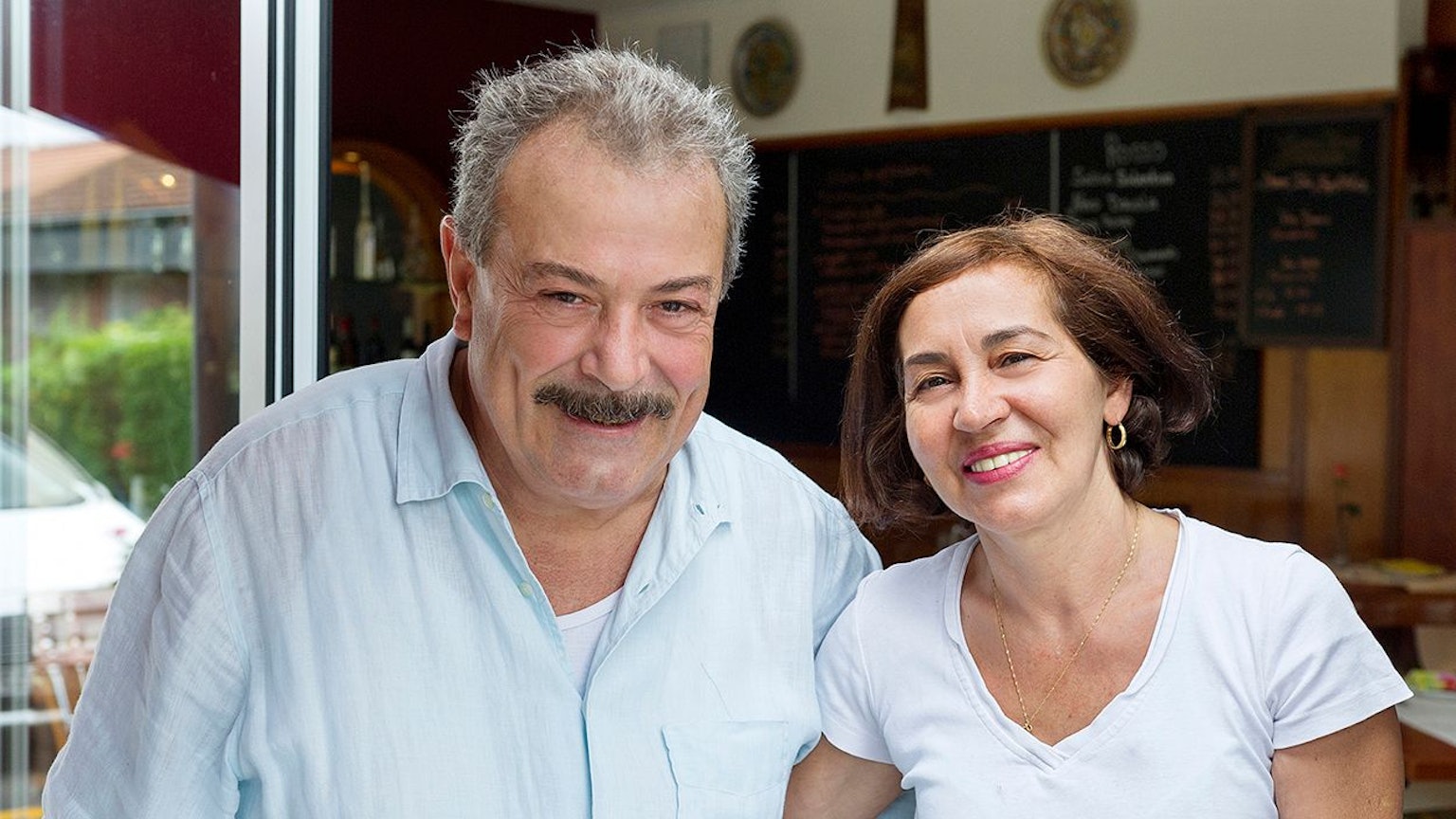 Ein Mann mit Schnauzer und eine Frau mit dunklem halblangem Haar schauen lächelnd in die Kamera.