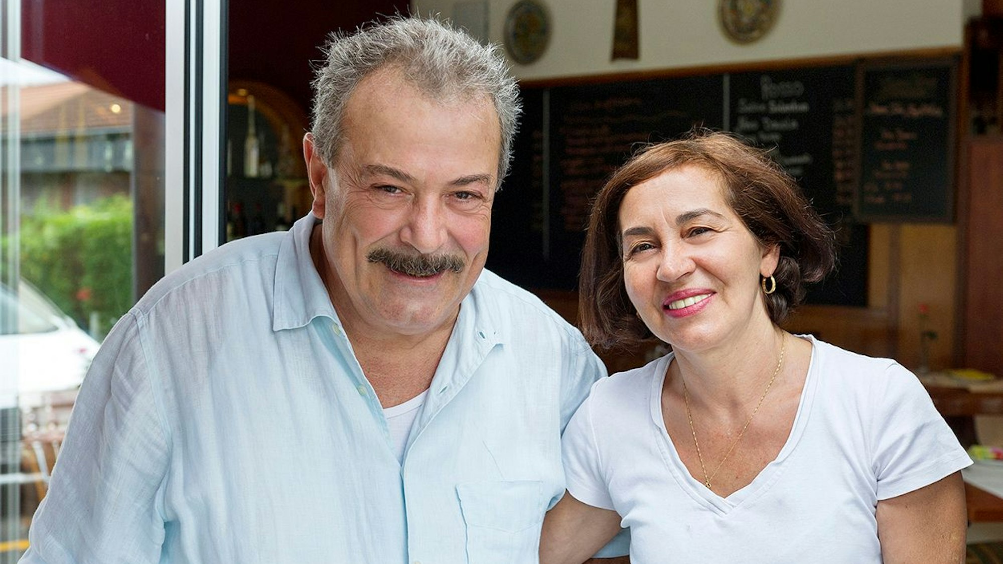 Ein Mann mit Schnauzer und eine Frau mit dunklem halblangem Haar schauen lächelnd in die Kamera.