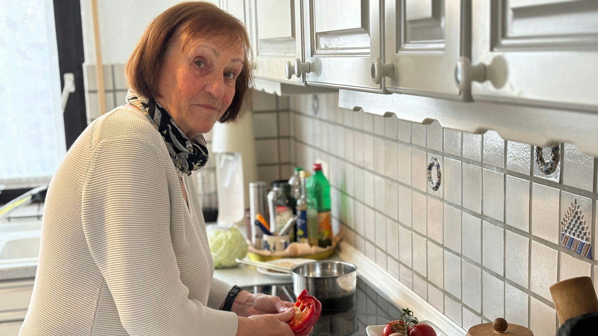 Eine Frau mit kinnlangem Haar, die Pullover und Schal trägt, steht in einer Küche und schneidet eine Paprika.