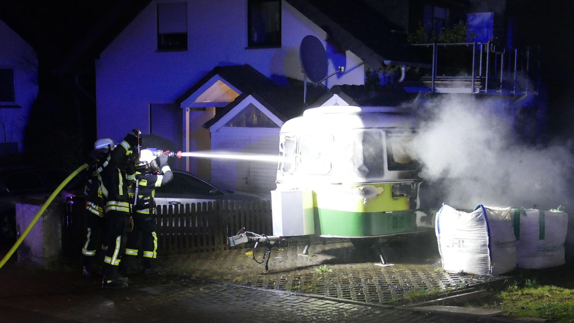 Feuerwehrleute richten einen Schlauch auf einen brennenden Wohnwagen.