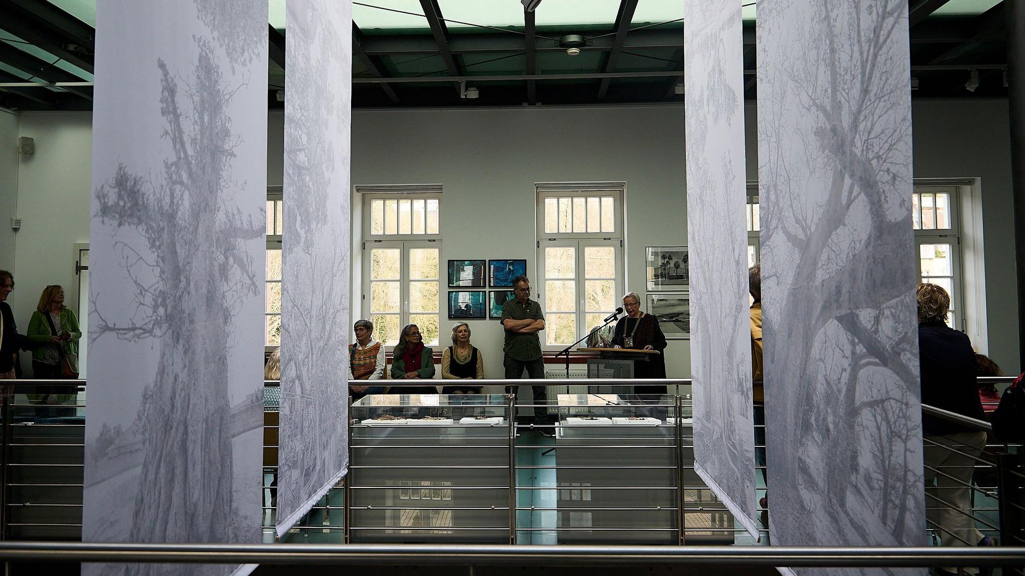 Fotografien von Rendel Freude hängen in der Mitte des Kunstforums. Im hinteren Bereich sind Menschen zu sehen. Eva-Maria Hermanns steht an einem Pult und spricht in ein Mikrofon.