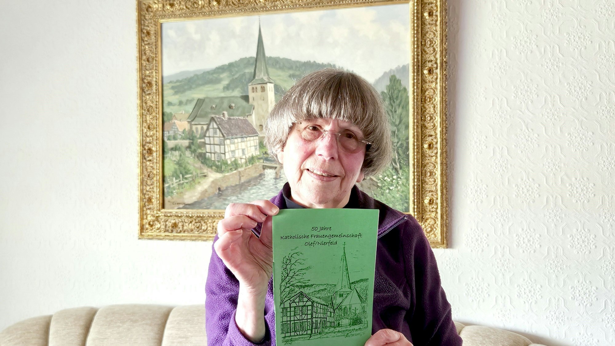 Marga Käßbach sitzt vor einem Gemälde mit der Olefer Pfarrkirche und hält die alte Festschrift.in beiden Händen.