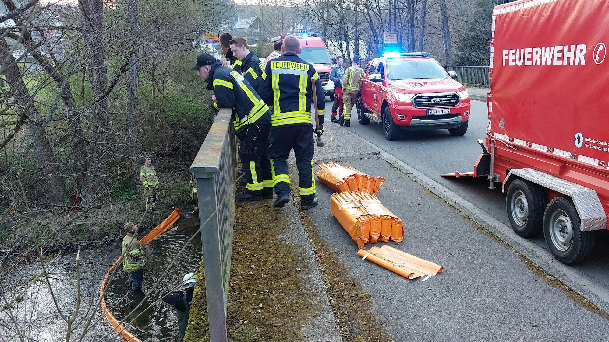 Feuerwehrleute auf einer Brücke und in einem Fluss. Orangefarbene Ölsperren im Einsatz.