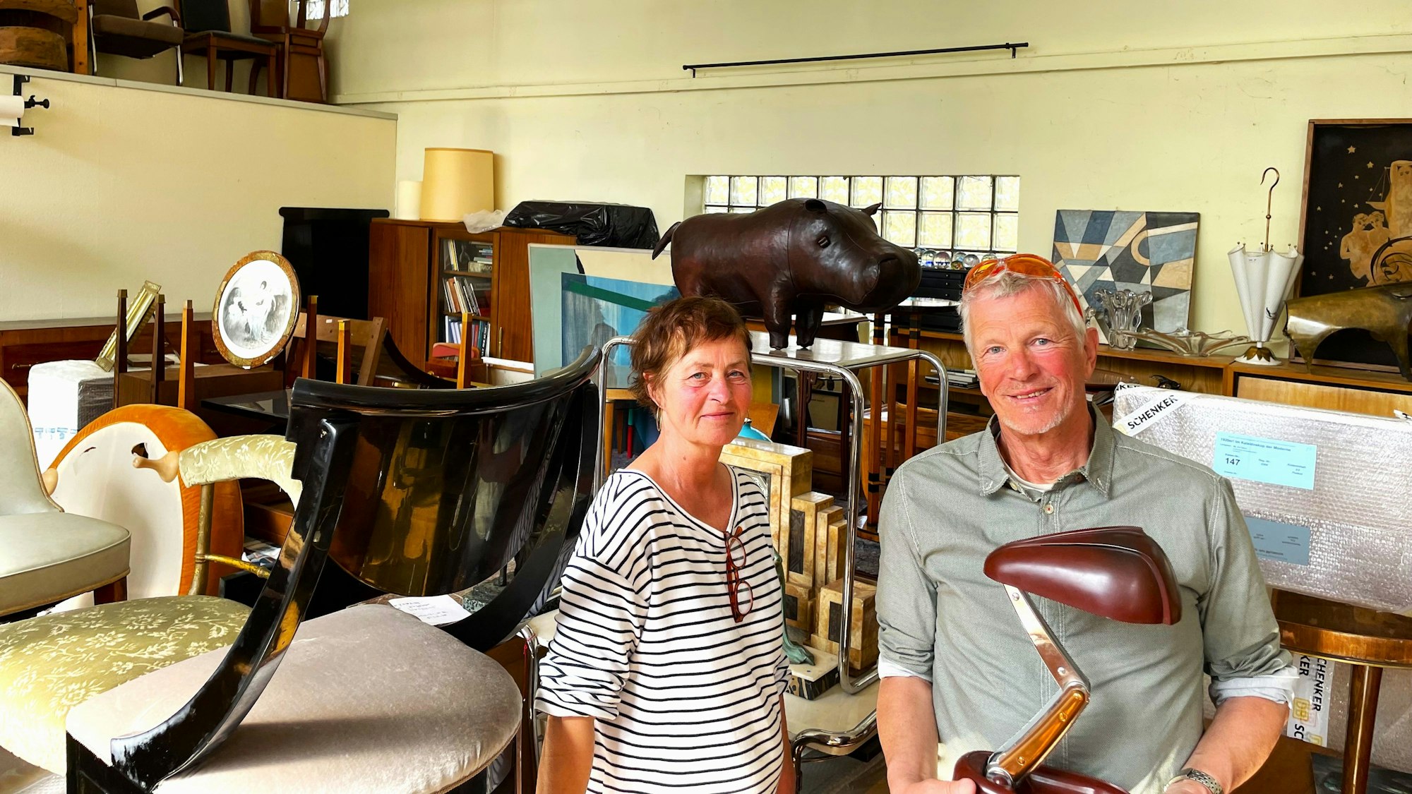 Eine Frau im Ringelshirt und ein Mann mit grauem Haar stehen in einem Lagerraum voller Möbel und Deko-Objekte.
