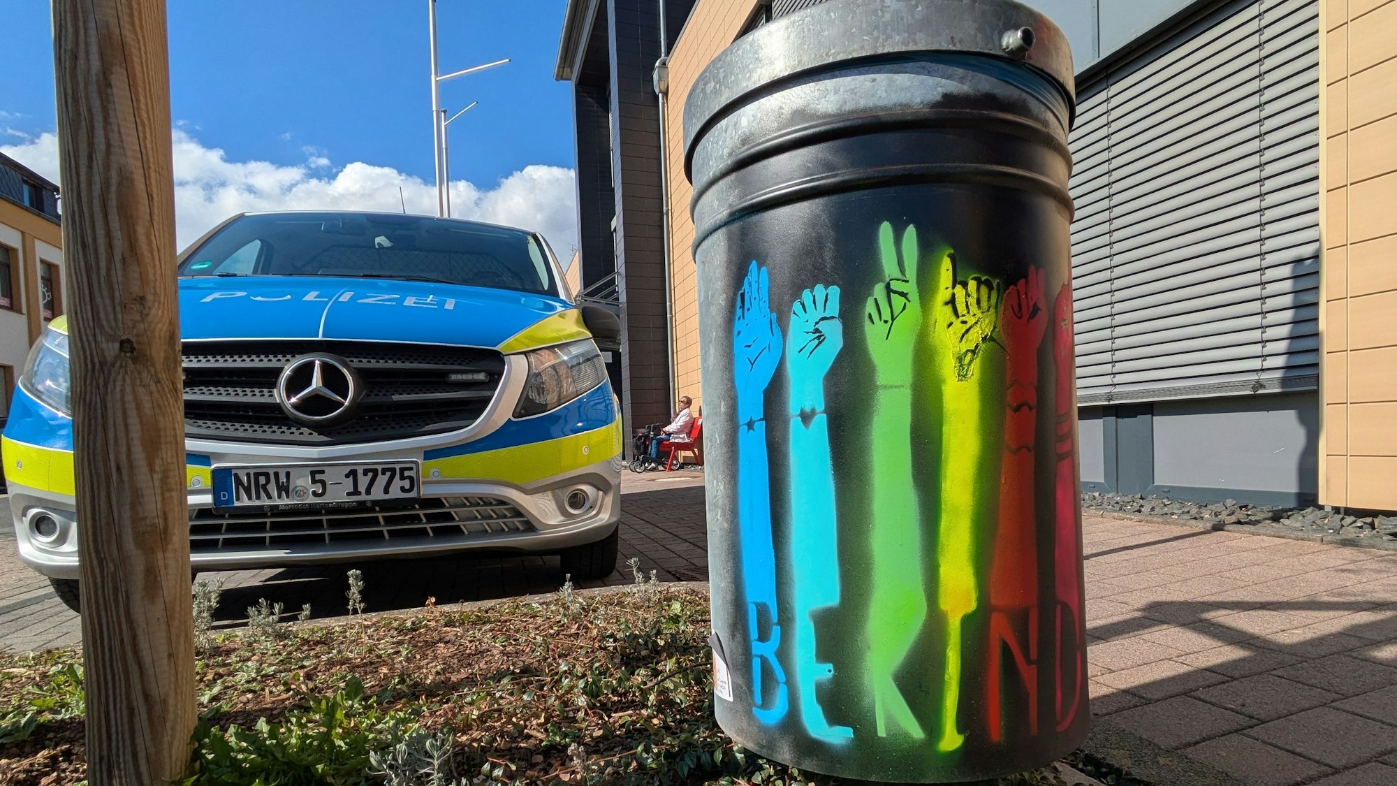 Der englischsprachige Schriftzug „Be kind“ ist auf einer in verschiedenen Farben besprühten Mülltonne zu lesen. Links steht ein Einsatzfahrzeug der Polizei.