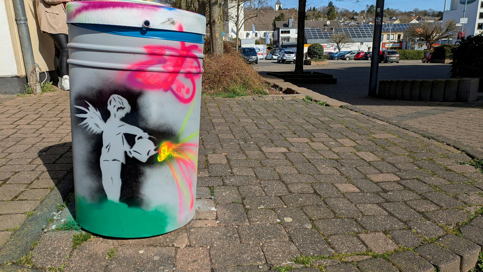 Auf einem Mülleimer aus grauem Zinkblech ist mit einer Schablone die Silhouette eines Engels mit einem Gießkännchen, aus dem sich bunte Farben ergießen, aufgesprüht worden.