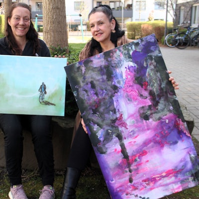 Zwei Frauen zeigen ihre Gemälde. Sie sitzen auf einem Baumbeet.