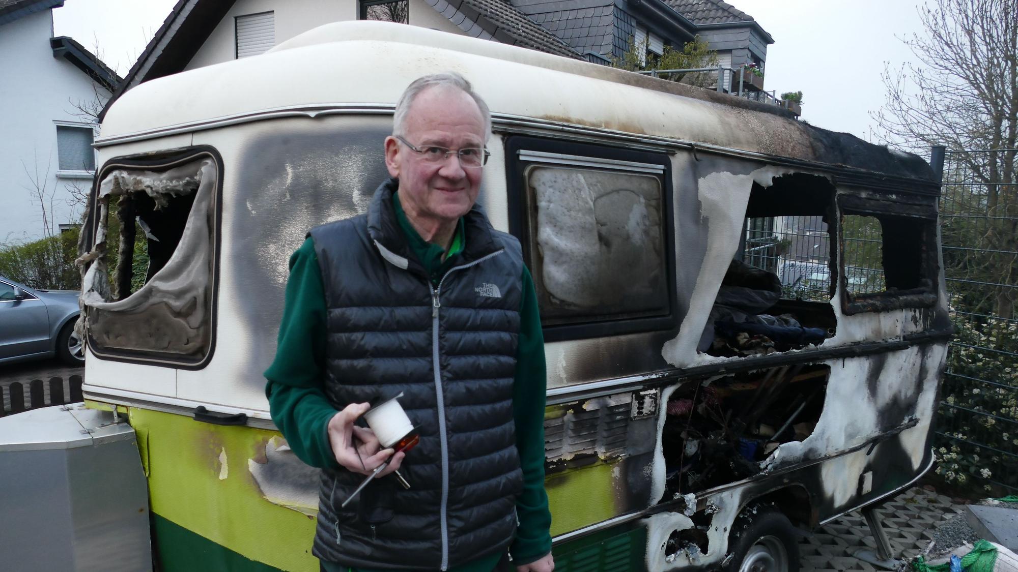 Ein Mann steht vor einem von Feuer völlig zestörten Wohnwagen.