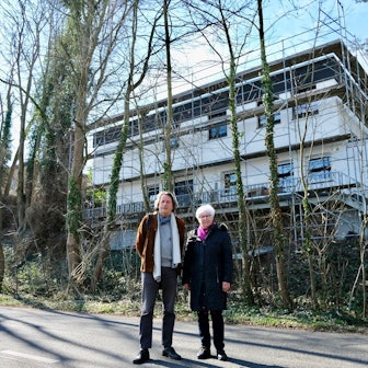 Ein Mann und eine Frau stehen vor einer Hangkante und einem Neubau. Vor dem Neubau stehen Bäume, die gefällt werden sollten.