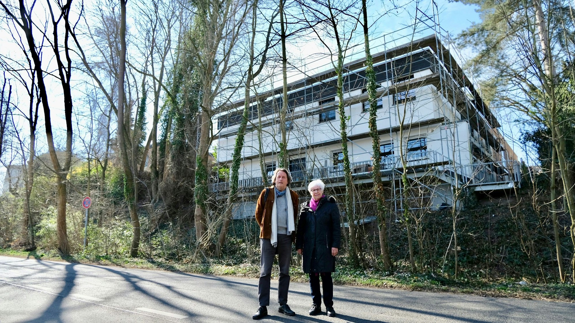 Ein Mann und eine Frau stehen vor einer Hangkante und einem Neubau. Vor dem Neubau stehen Bäume, die gefällt werden sollten.