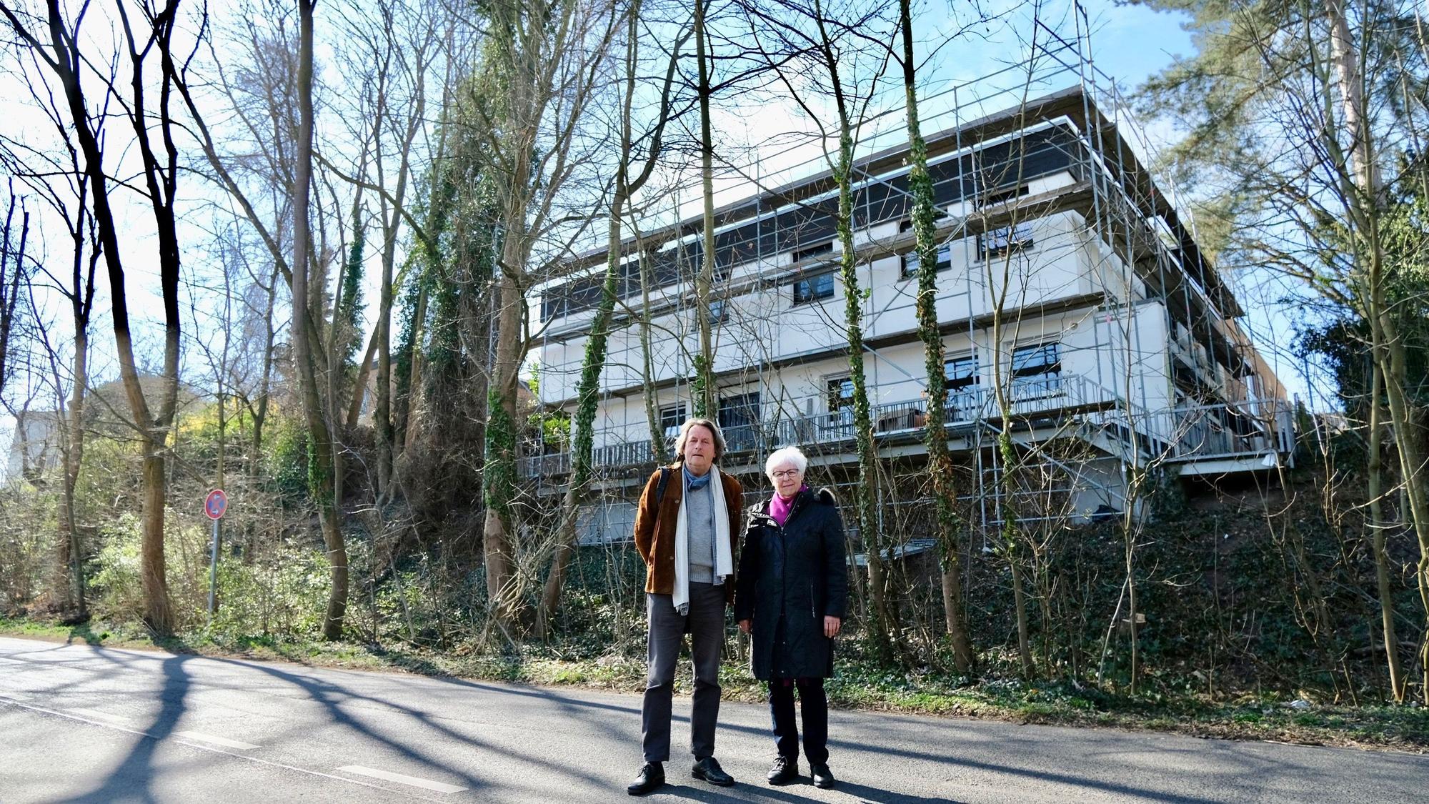 Ein Mann und eine Frau stehen vor einer Hangkante und einem Neubau. Vor dem Neubau stehen Bäume, die gefällt werden sollten.
