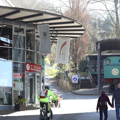 Die Glasfassade am Eingang der Drachenfelsbahn, im Hintergrund eine grüne Lok, die als Denkmal installiert ist.